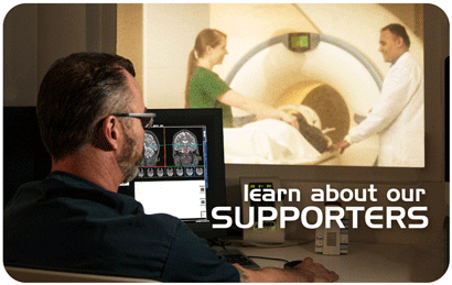 Photo of Technologist in MR Control room with scanner, researchers and participant visible in the background